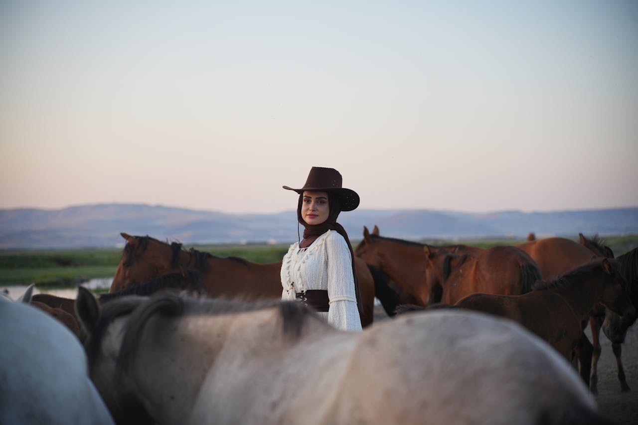 Home A cowgirl in a brown hat stands among horses in a vast, scenic landscape with distant hills.