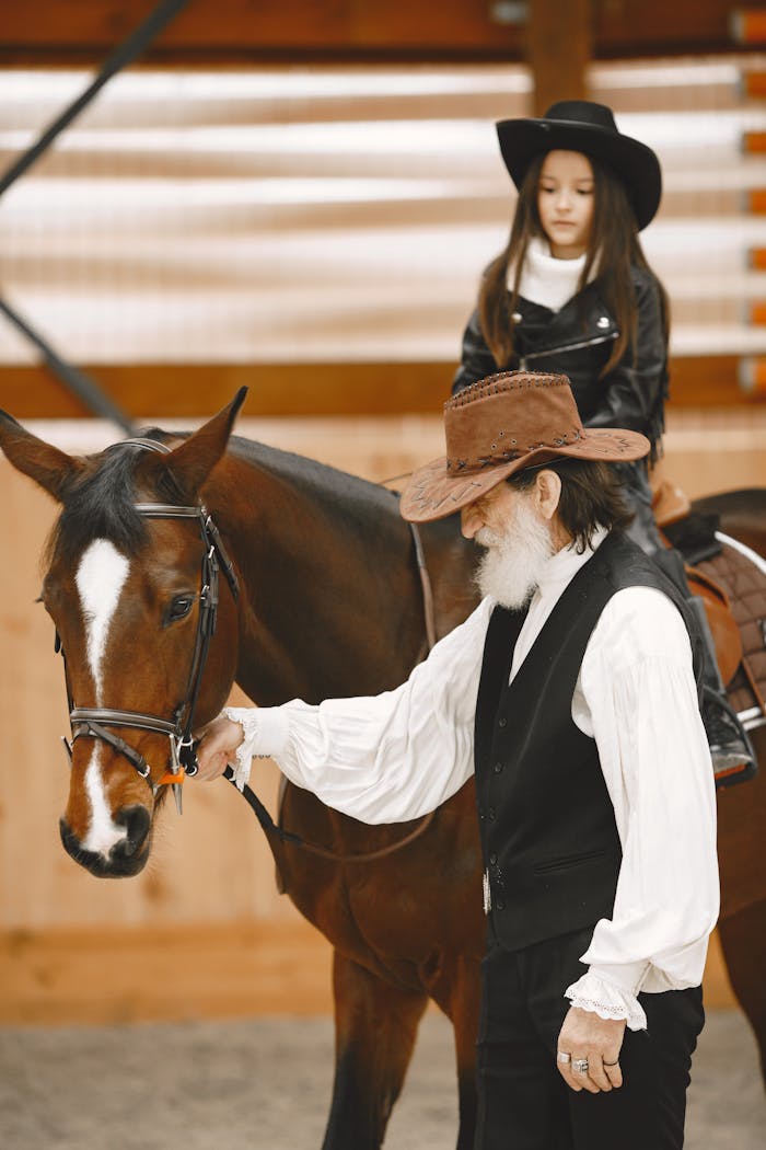 Home An elderly cowboy assists a young cowgirl riding a brown horse indoors, showcasing equestrian skills.